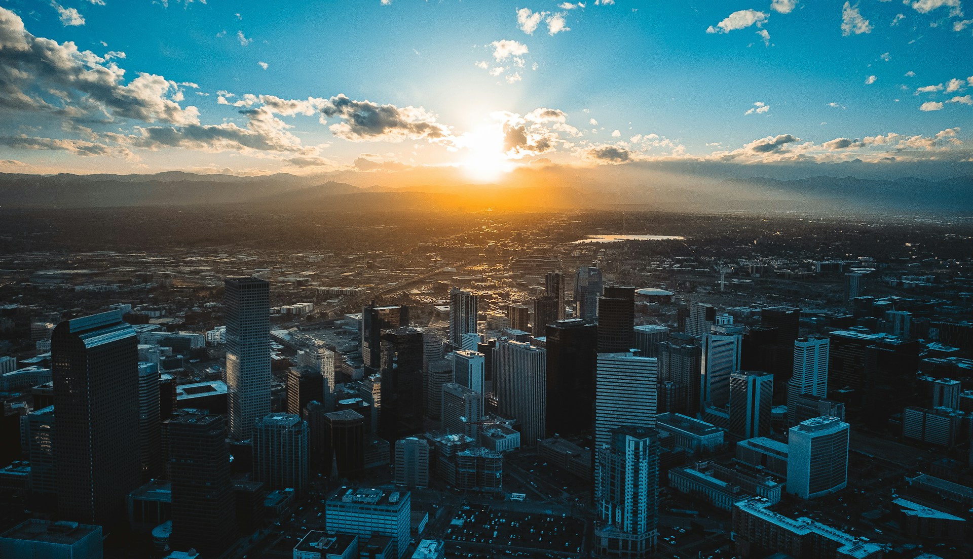 Aerial view of downtown Denver, Colorado at sunset, with sunlight casting long shadows over skyscrapers and the Rocky Mountains visible in the distance.