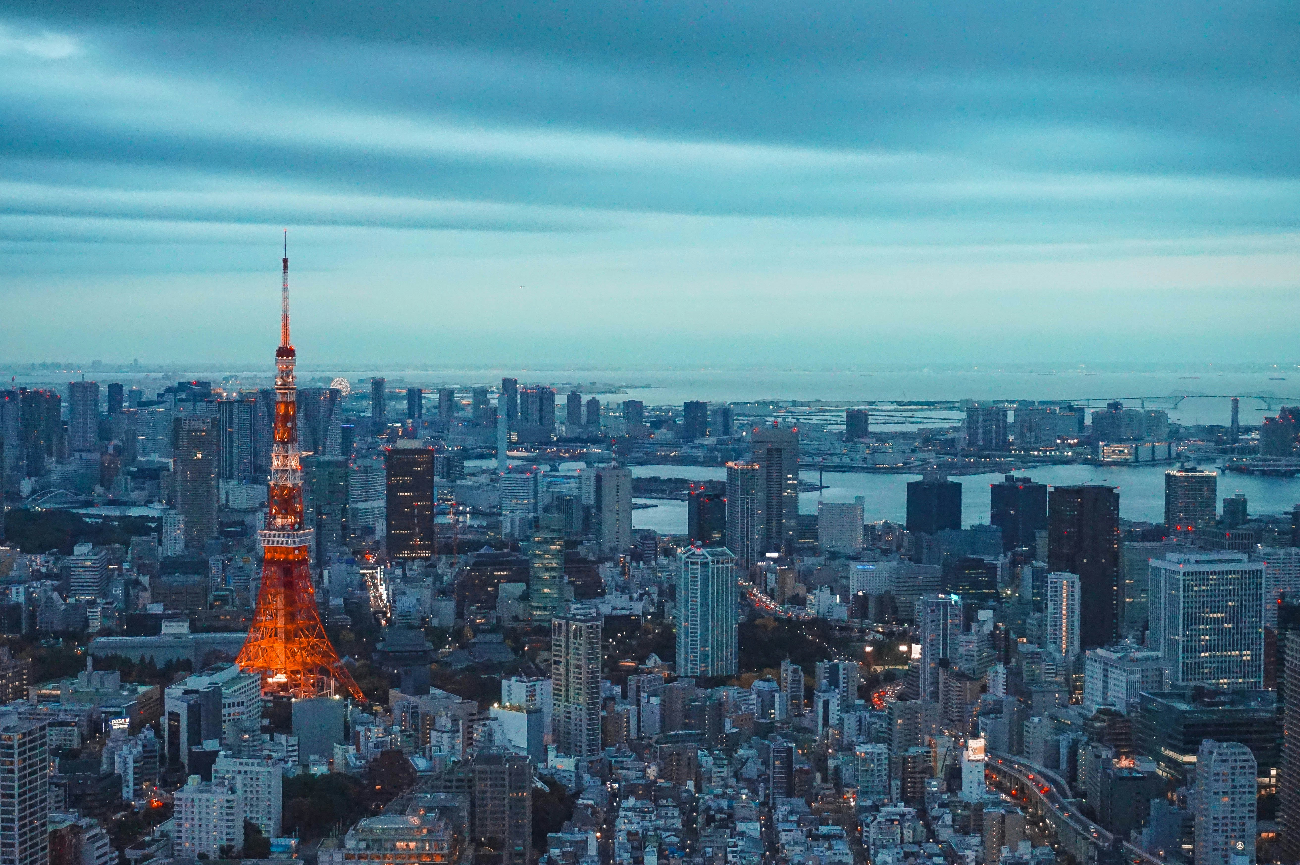 Aerial view of Tokyo at dusk, featuring Tokyo Tower illuminated in orange-red lights, surrounded by dense urban high-rises and the Tokyo Bay waterfront under a cloudy blue sky.