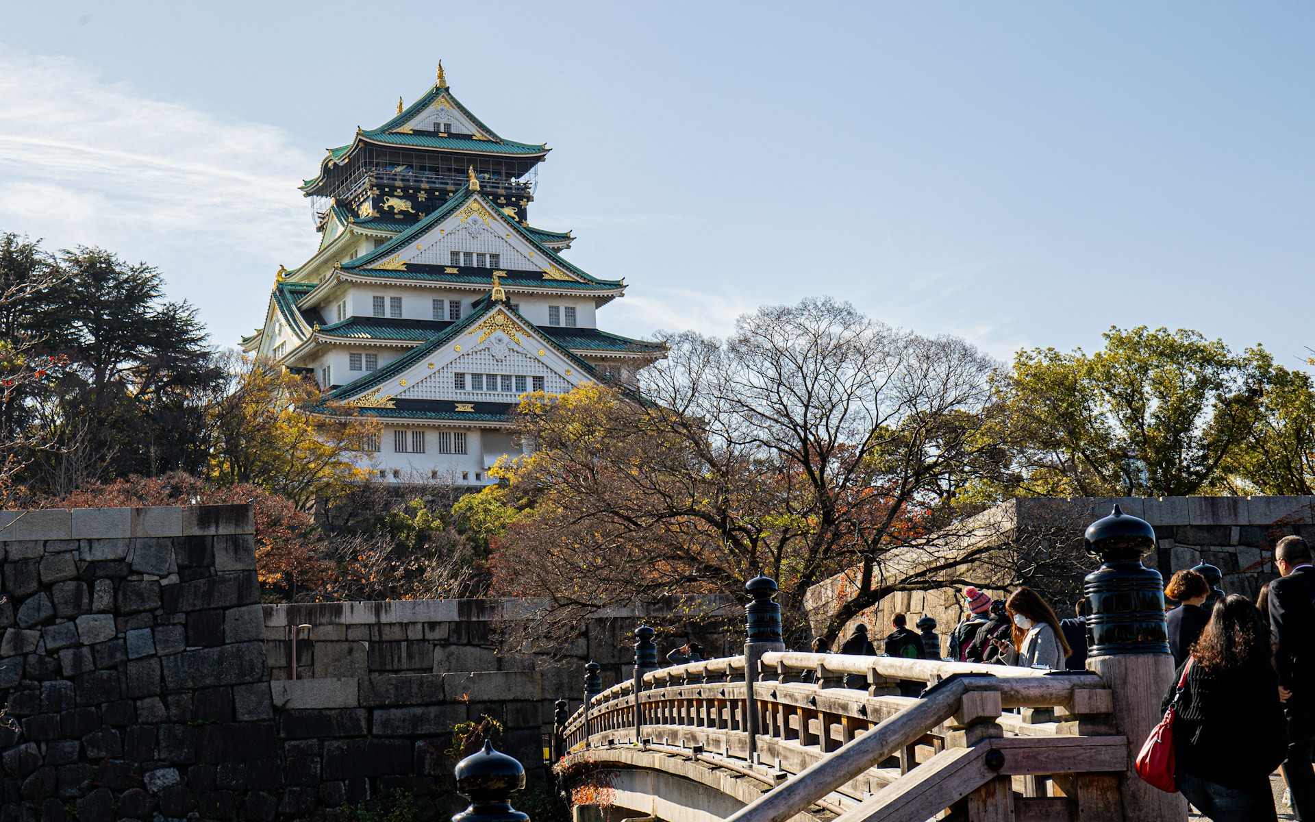 View of Osaka Castle with its green rooftops and gold accents, surrounded by autumn trees, as visitors cross a wooden bridge in the foreground under a clear blue sky.