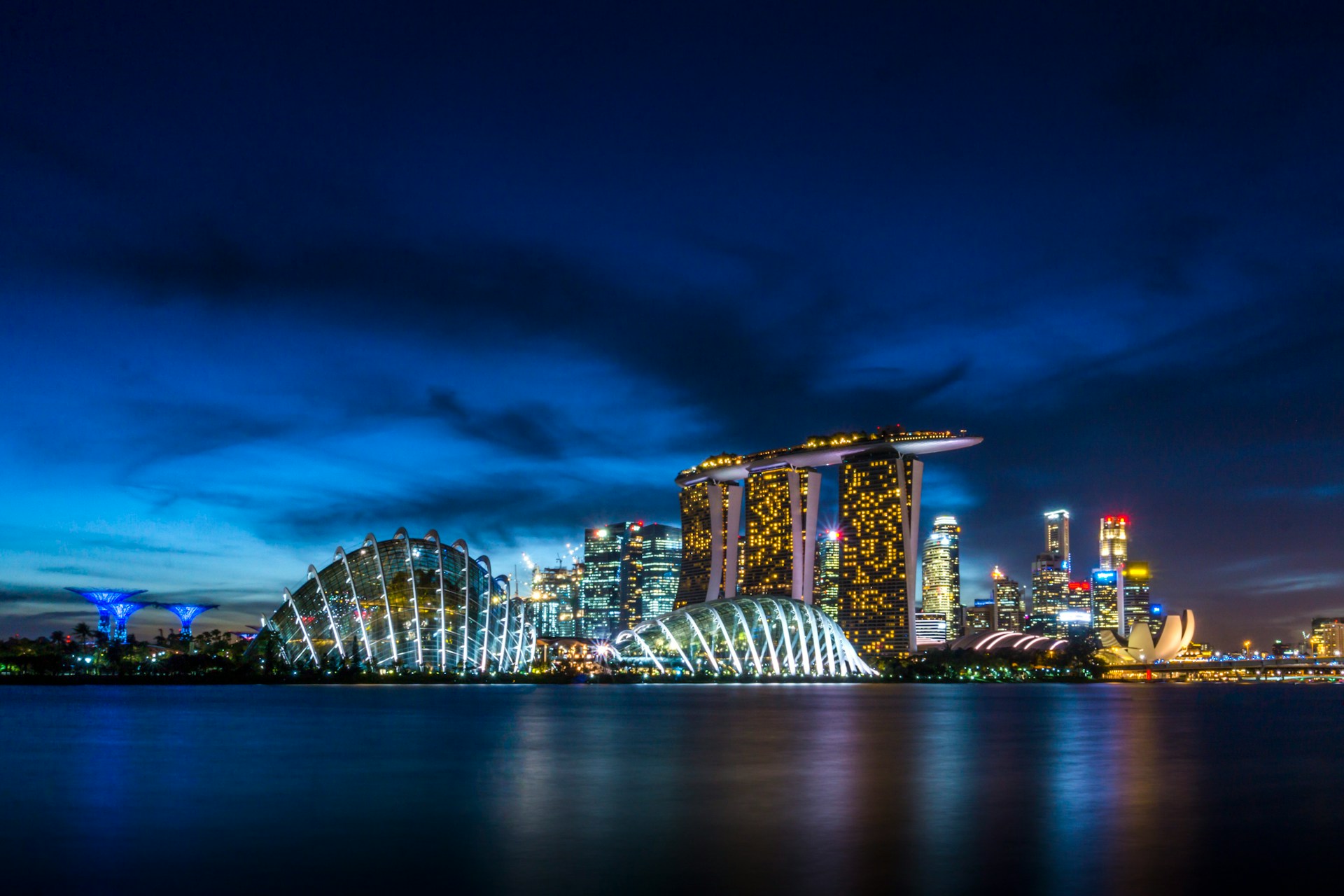 Singapore skyline at night, featuring Marina Bay Sands, Supertree Grove, and illuminated domes of Gardens by the Bay reflected in calm water.