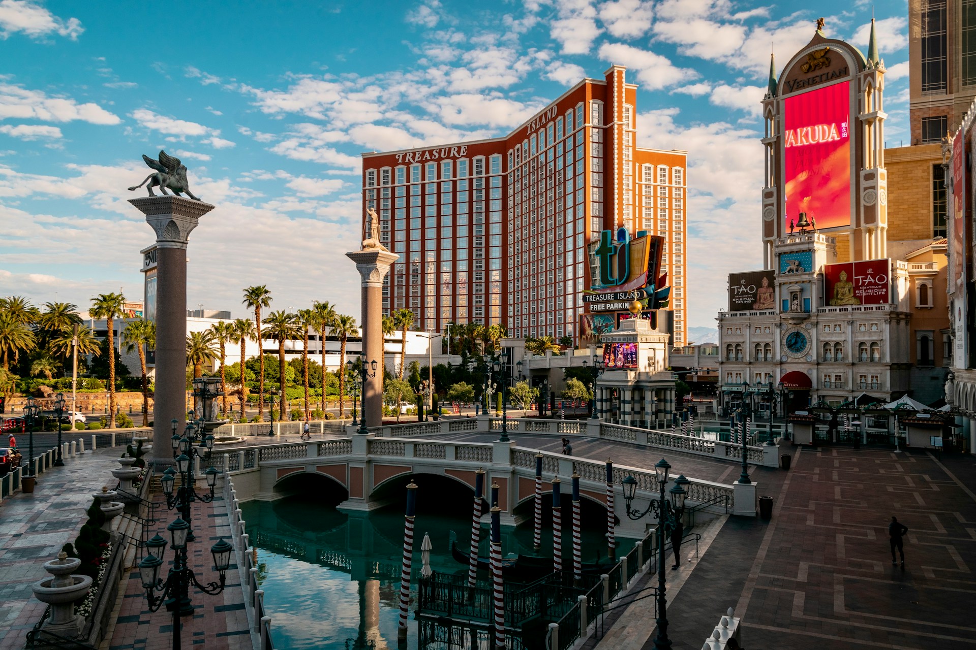 The exterior of The Venetian resort on the Las Vegas Strip, featuring a replica Venetian canal with a arched bridge, gondola posts, and ornate lampposts in the foreground. The Treasure Island hotel tower rises in the background, with The Venetian's facade — including its iconic winged lion columns and TAO restaurant signage — visible on the right, under a partly cloudy blue sky.