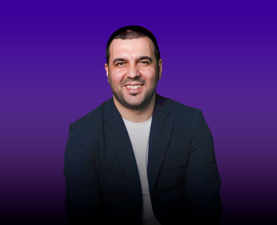 Mor degrade arka plan önünde koyu ceket ve açık renk tişört giymiş, gülümseyen erkek portresi.