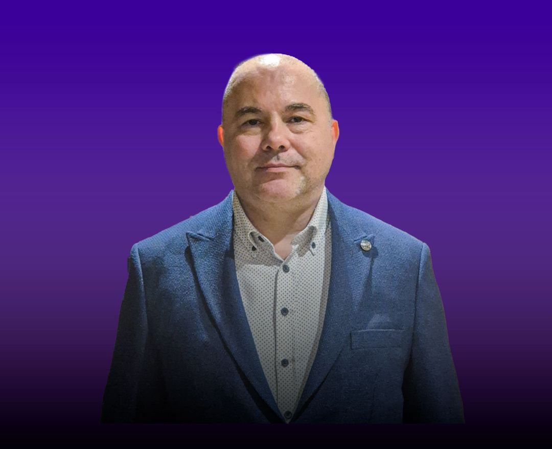Mor arka plan önünde mavi ceket ve gömlek giymiş orta yaşlı bir erkek.
