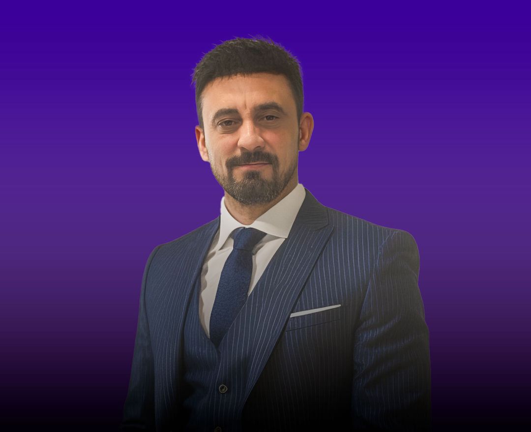 Mor arka plan önünde koyu takım elbise ve kravat giymiş sakallı bir erkek.