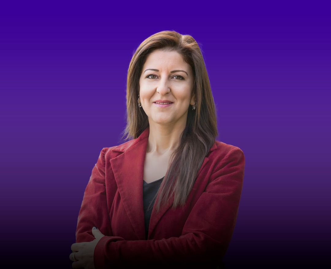 Kırmızı ceket giymiş, uzun saçlı bir kadın mor degrade arka plan önünde kollarını çaprazlayarak kameraya gülümsüyor.