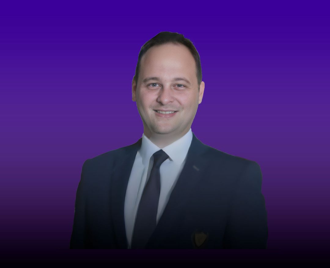 Takım elbise ve kravat giymiş bir adam, mor degrade arka plan önünde gülümseyerek kameraya bakıyor.