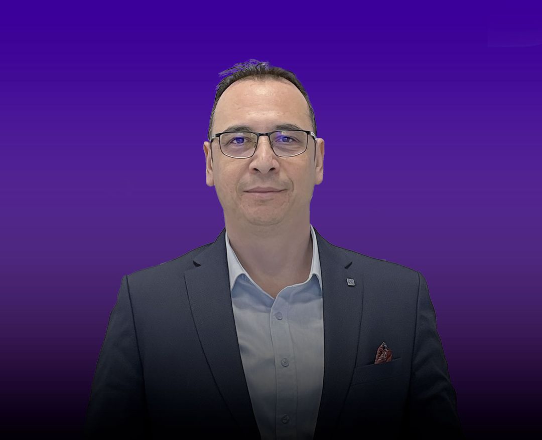 Gözlüklü, koyu renk bir ceket giymiş bir adam, mor tonlu degrade arka plan önünde kameraya bakıyor.