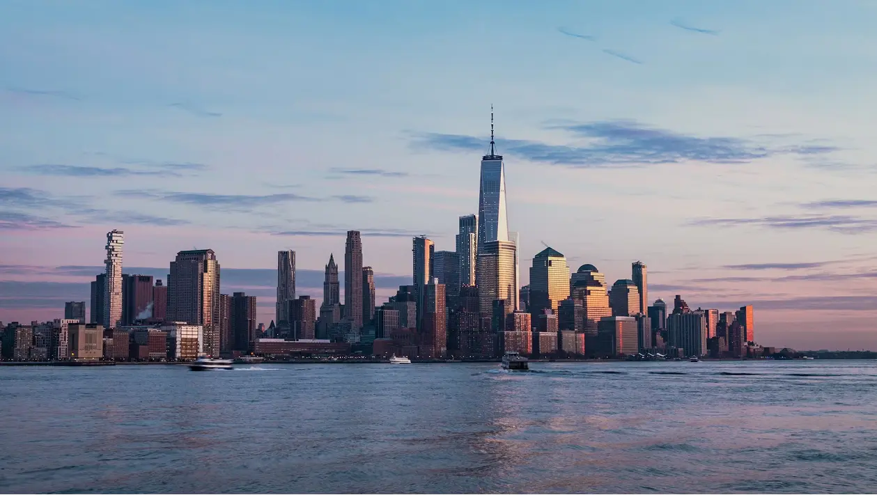 New York City skyline at sunset with One World Trade Center towering over buildings by the water.