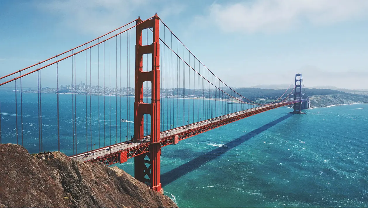 Golden Gate Bridge spanning blue water with city skyline and hills in the background on a clear day.