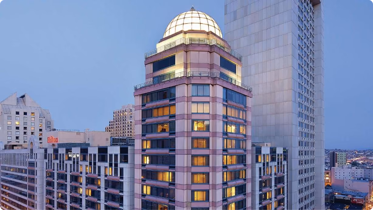 Exterior photo of a hotel tower with a domed rooftop lit up at dusk, part of a city skyline