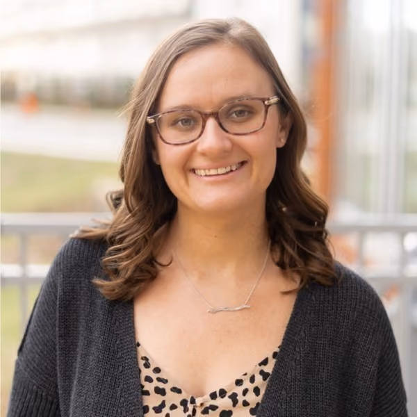 Smiling woman with shoulder-length curly brown hair, wearing glasses, a leopard print top, a dark cardigan, and a necklace, standing outdoors with blurred background.