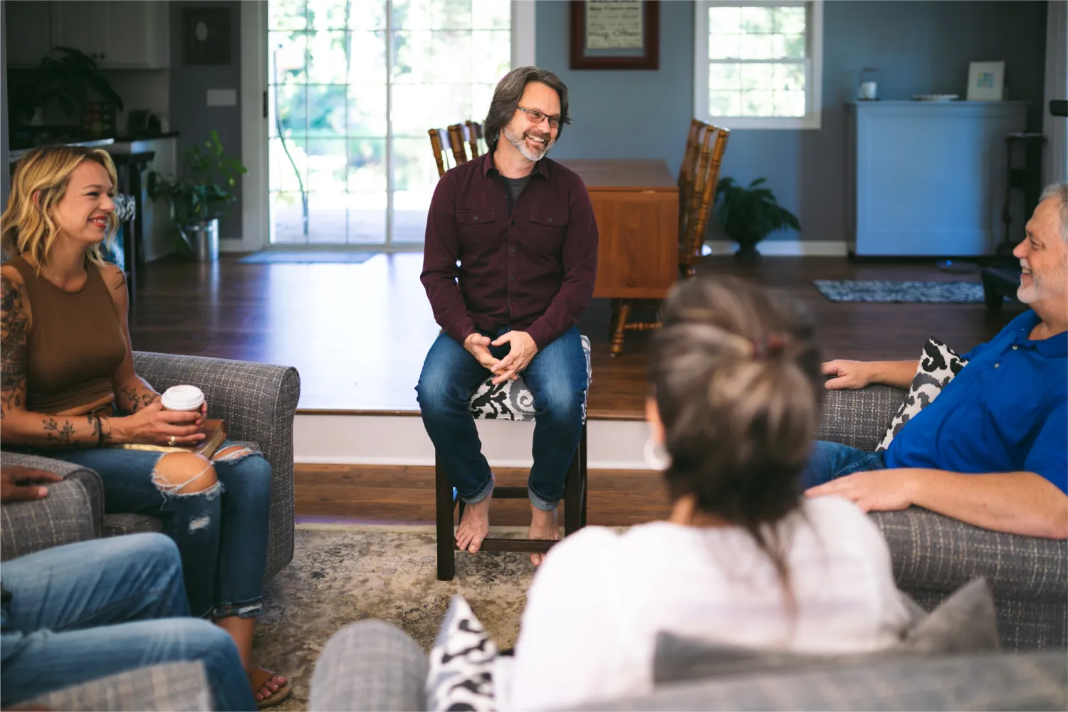 Chuck Coward (The Struggle Coach) seated on a stool smiling and speaking with a group of people in a living room setting.