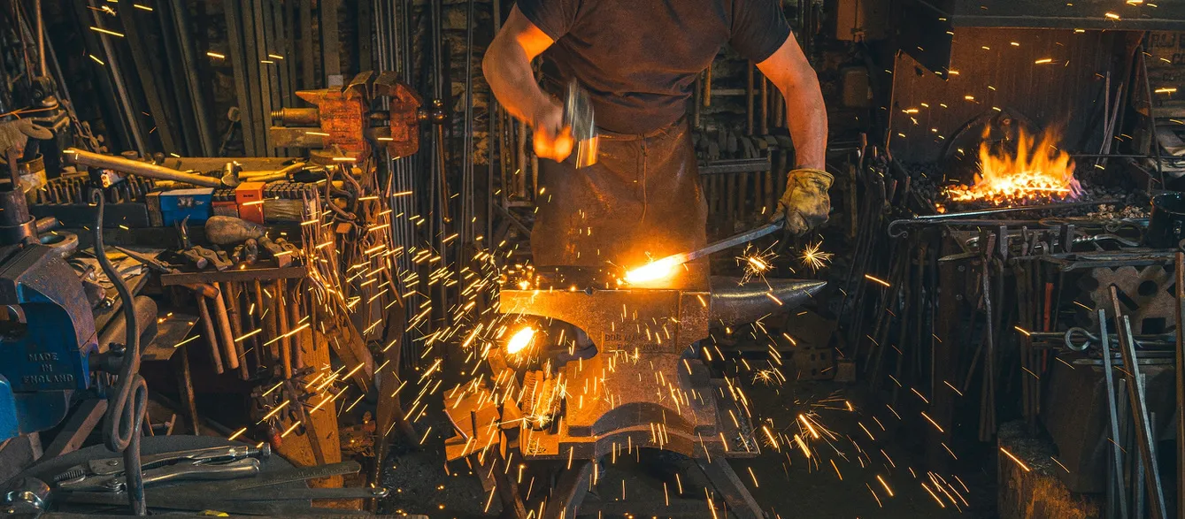A blacksmith surrounded by sparks and tools works in a busy, fiery workshop.
