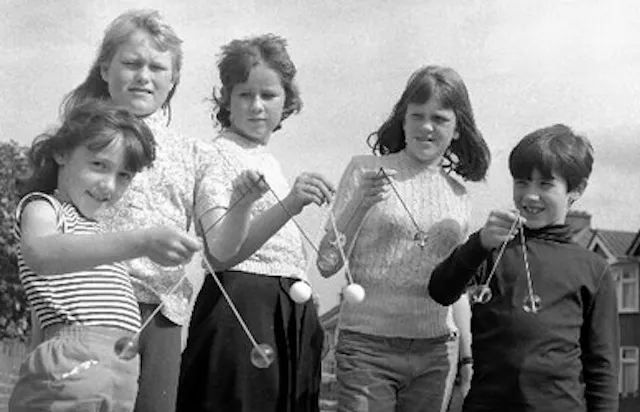 Vintage black-and-white photo of a group of children playing outside with Clackers, a toy made of two hard balls on strings that click together when swung.