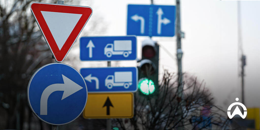 A collection of road signs including a yield sign, right turn arrow, truck direction signs, and a green traffic light.