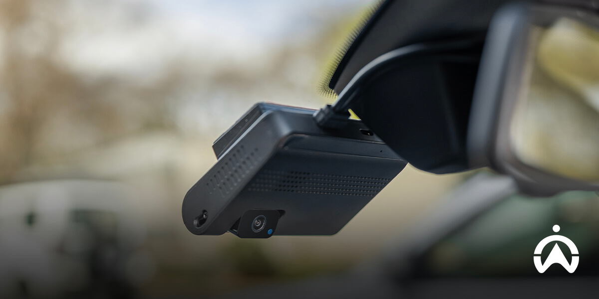 A close-up of a black dashcam mounted on a car's windshield, with a blurred background of trees and vehicles.