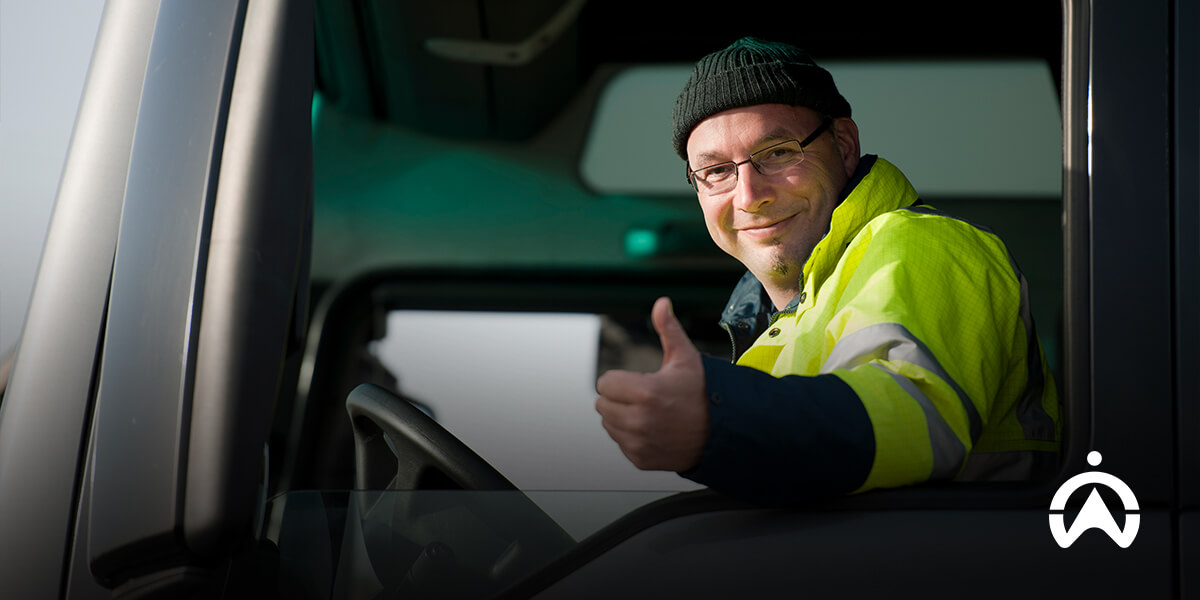 A truck driver in a bright safety jacket smiles and gives a thumbs-up from the window of his vehicle.