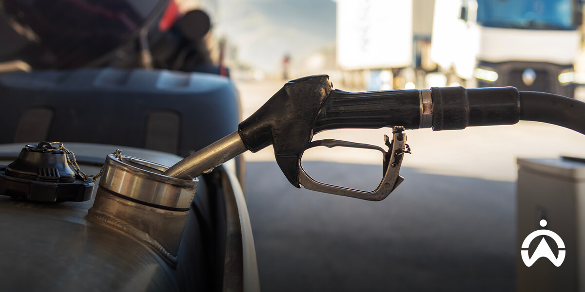 Close-up of a fuel nozzle being inserted into a vehicle's tank, with blurred trucks in the background at a gas station.