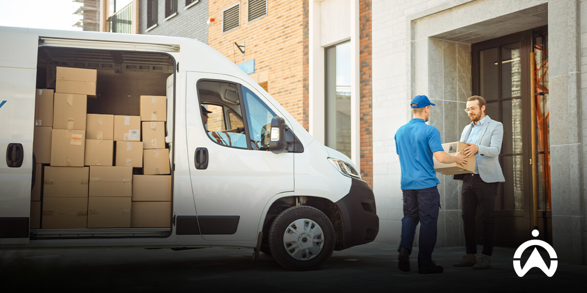 A delivery person hands a package to a recipient outside a building, with a van loaded with cardboard boxes in the background.