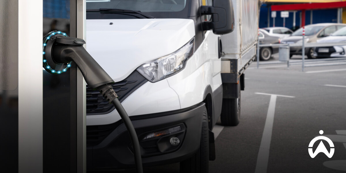 An electric vehicle charging connector plugged into a station, with a white delivery truck parked nearby.