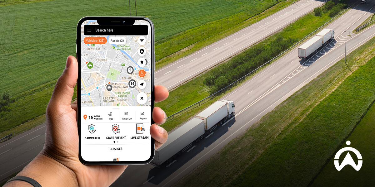 A hand holds a smartphone displaying a tracking app while trucks drive on a highway with greenery along the roadside.