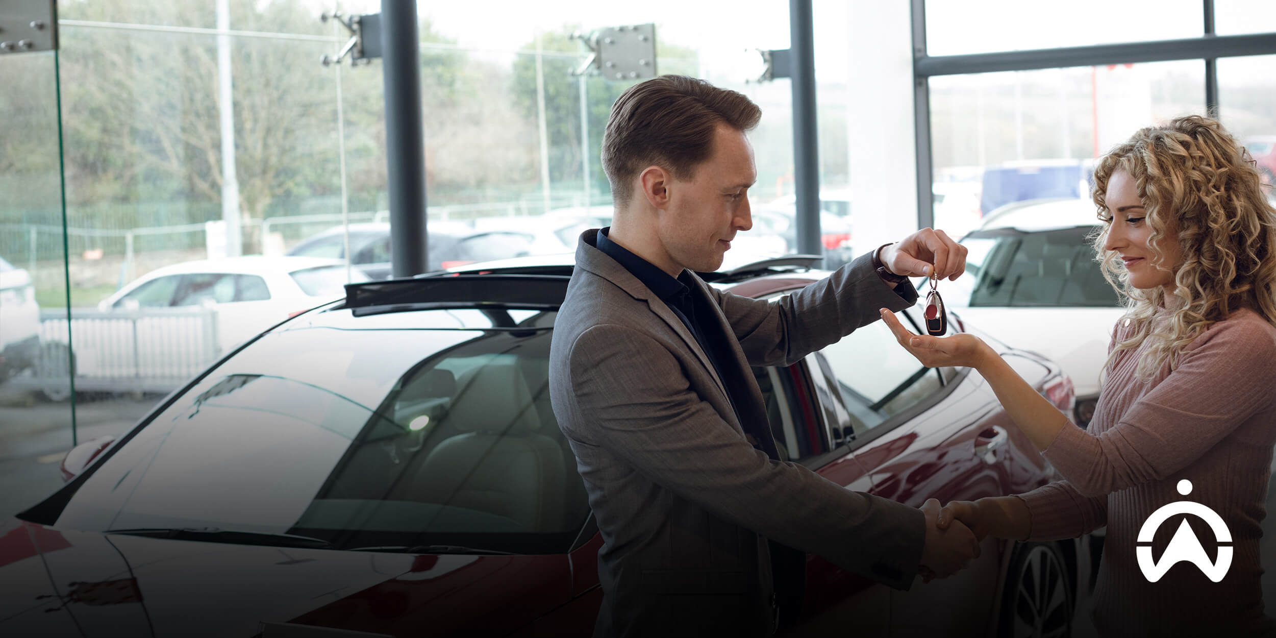 A man in a suit hands car keys to a woman in a dealership, with vehicles visible in the background.