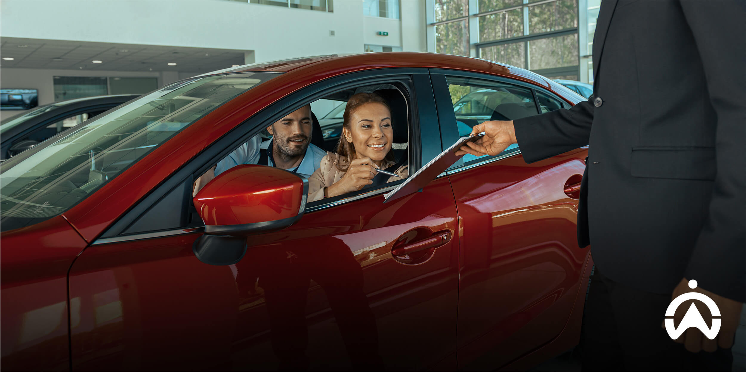 A customer in a red car is discussing details with a salesperson, who holds a document, inside a car dealership.