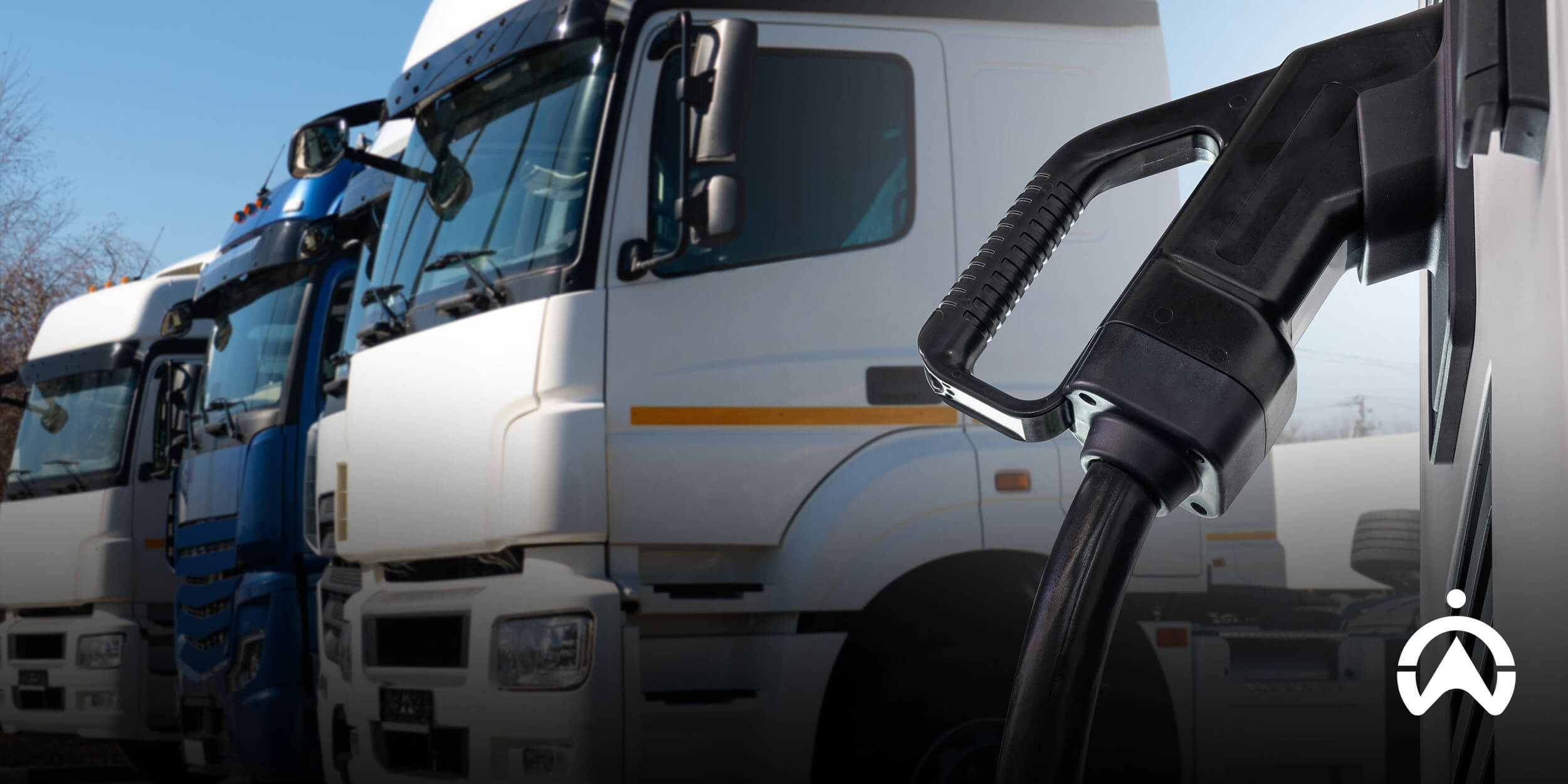 Close-up of a fuel nozzle in the foreground with multiple commercial trucks lined up under a clear blue sky, illustrating the transition phase within a fleet electrification roadmap from traditional fuel vehicles to electric fleets.