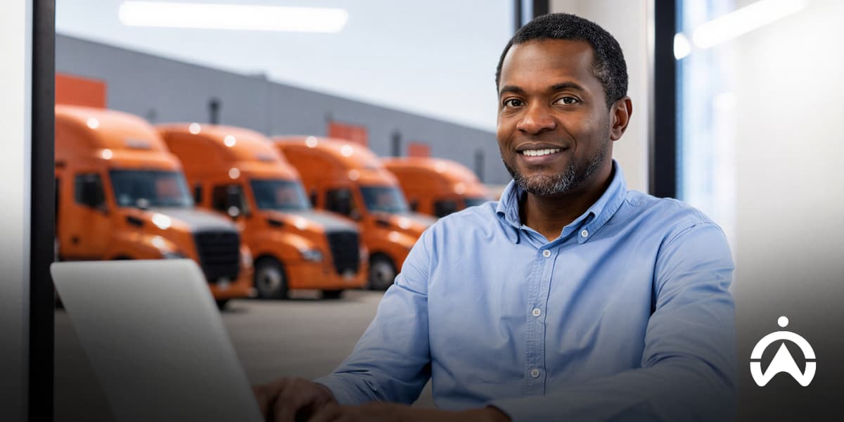 Smiling fleet manager using a laptop in front of orange semi-trucks, illustrating the Scopes of Carbon Tracking.