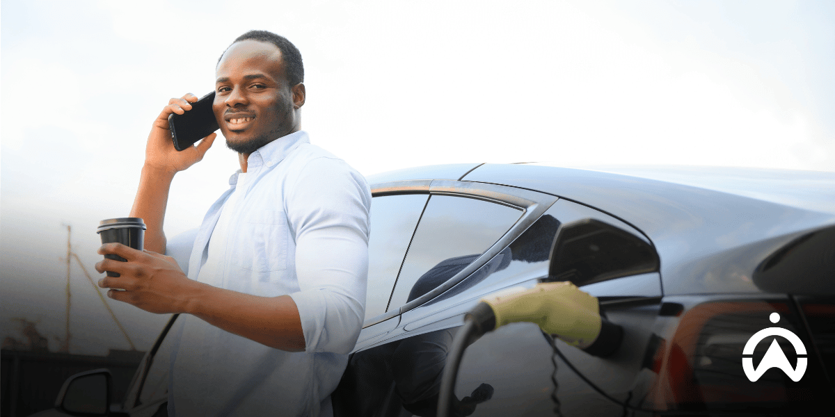 A man holding a coffee in one hand and in the other, a phone to his ear, whilst leaning against a black car with a charger plugged into it