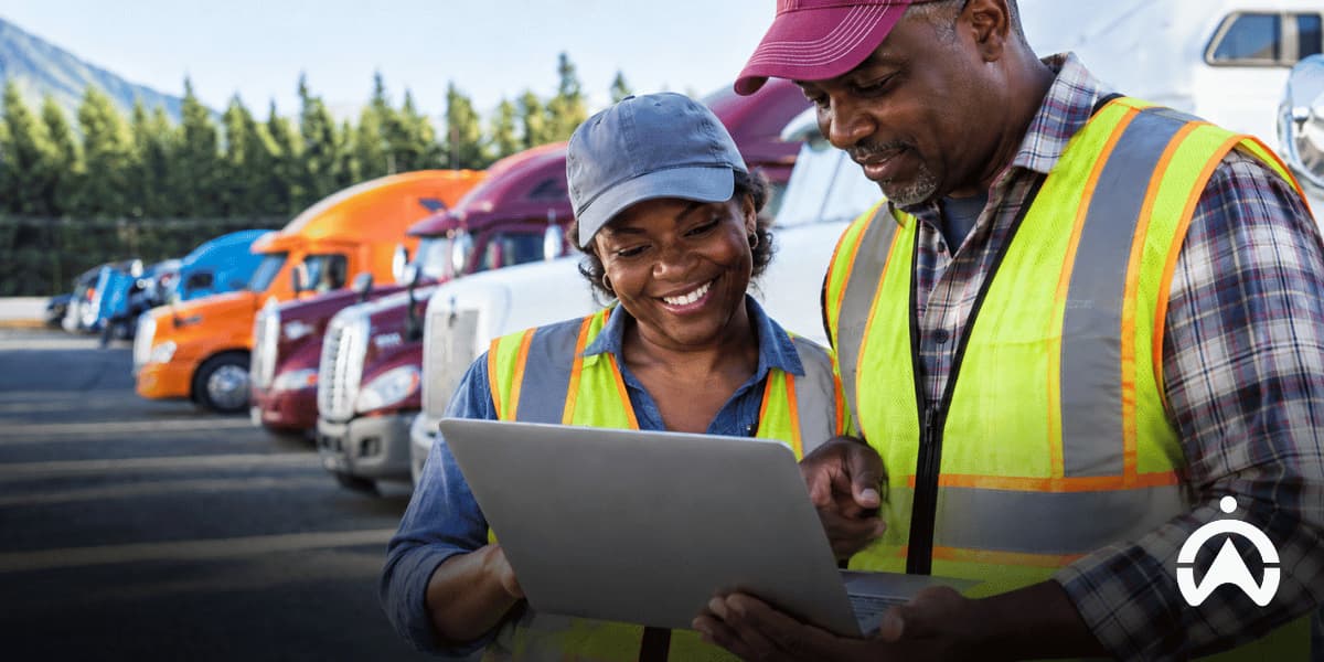 Two truck workers reviewing driver performance data on a laptop beside parked semitrucks to assess truck driver driving habits and safety.