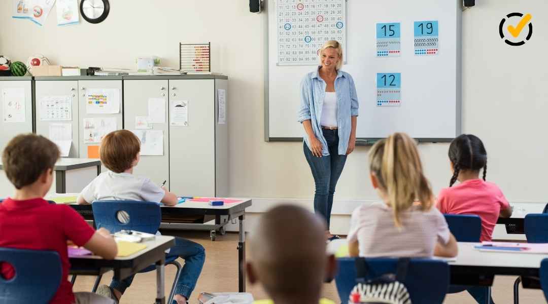 "A smiling female teacher stands at the front of a classroom, facing several elementary school students seated at desks. A white board behind her shows numbers and math visuals. The Formative by Newsela logo is in the top right corner."
