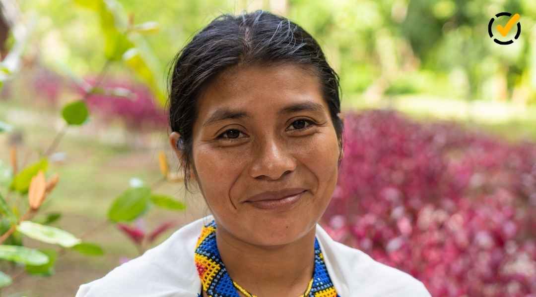 "A smiling Indigenous woman with dark hair and a colorful beaded necklace looks directly at the camera, against a blurred background of green and red foliage. The Formative by Newsela logo is in the top right corner." 