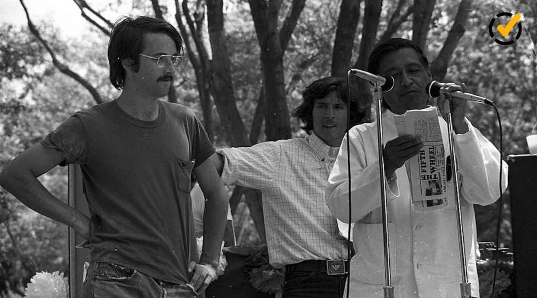 "A black and white photo of civil rights leader Cesar Chavez speaking into microphones, holding a newspaper titled 'The Fifth Wheel'. Two younger men stand behind him. The Formative by Newsela logo is in the top right corner."