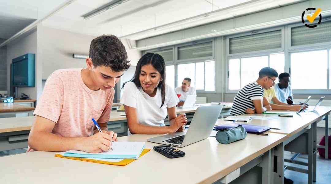 "Two students, a young man and a young woman, are working at a long desk in a classroom. The young man is writing in a notebook, while the young woman looks at a laptop. Other students are visible in the background. The Formative by Newsela logo is in the 