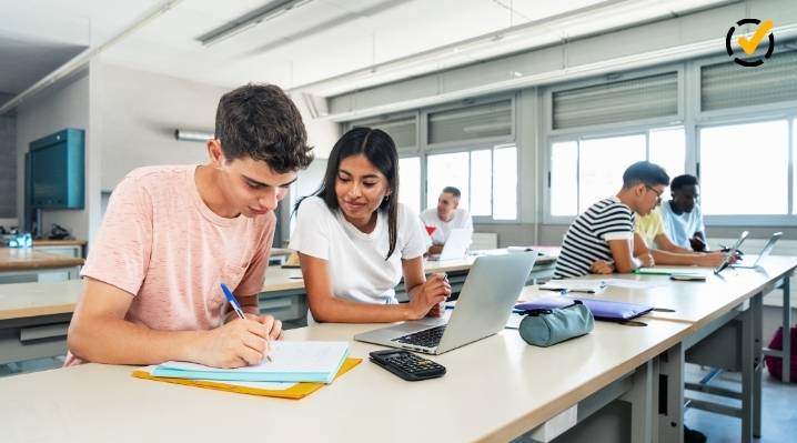 "Two students, a young man and a young woman, are working at a long desk in a classroom. The young man is writing in a notebook, while the young woman looks at a laptop. Other students are visible in the background. The Formative by Newsela logo is in the 