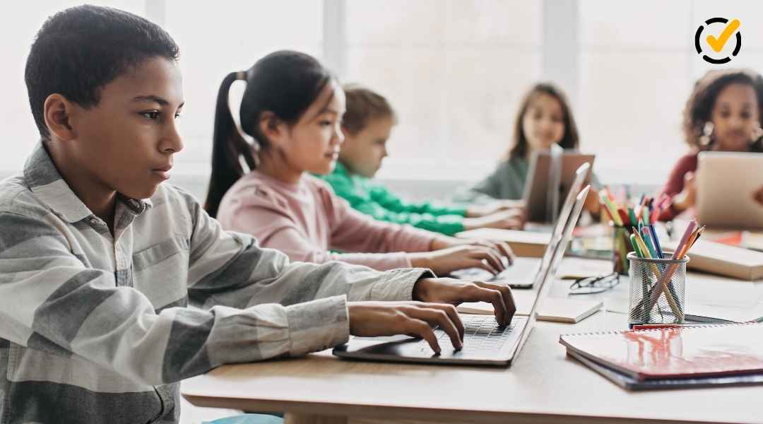 "A group of diverse elementary school children are seated at a long table in a bright classroom, actively engaged with laptops. School supplies like pens and notebooks are on the table. The Formative by Newsela logo is in the top right corner." 