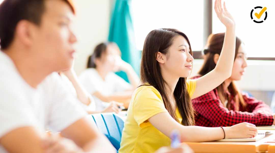 "A young female Asian student in a yellow shirt raises her hand in a classroom, with other students and a blurred background. The Formative by Newsela logo is in the top right corner."