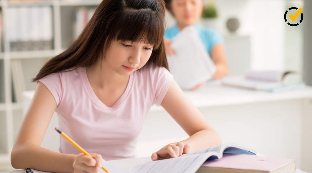 "A young female student with long dark hair and a pink shirt is writing in a notebook, with an open book beside her on a desk. Another student is visible, blurred, in the background. The Formative by Newsela logo is in the top right corner."