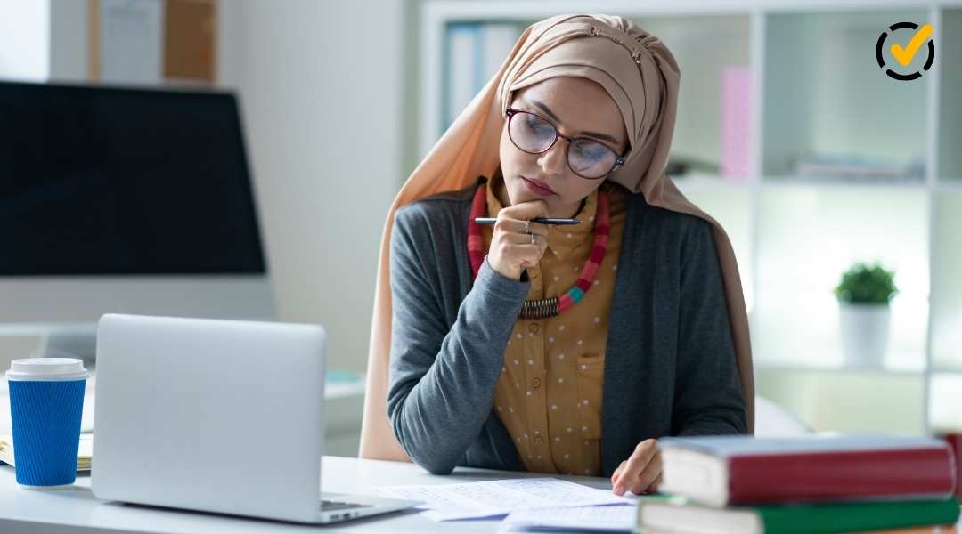 A teacher wearing a hijab, glasses, and a colorful necklace is seated at a desk, looking thoughtfully at a document while holding a pen to her chin. A laptop and stacked books are on the desk.