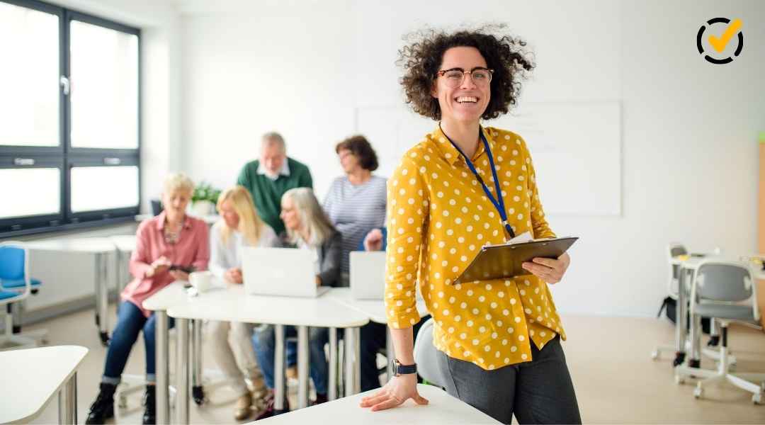 A smiling, curly-haired female teacher wearing a yellow polka-dot shirt stands in the foreground, holding a clipboard. In the blurred background, a diverse group of teachers are gathered around a table working on laptops, suggesting a Professional Learning