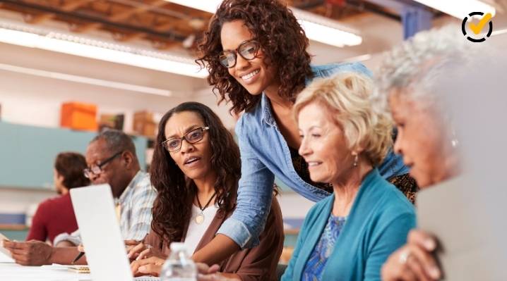 A diverse group of educators, primarily women, are collaborating around a laptop in a brightly lit professional setting. A younger teacher is standing and smiling as she helps or interacts with two older colleagues seated at the table.