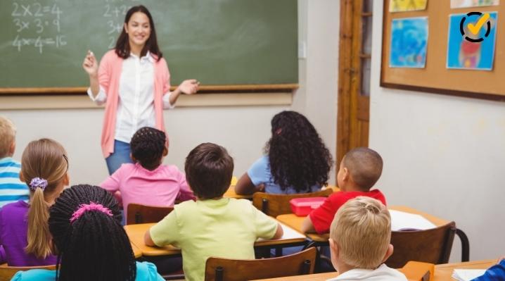 A smiling female elementary school teacher is standing in front of a classroom chalkboard, teaching a lesson to a diverse group of students seated at their desks, demonstrating the "I Do" phase of instruction.