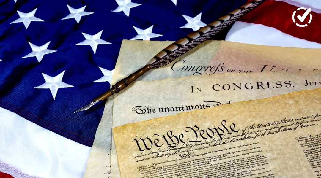 A close-up view of a decorative quill pen resting on aged historical documents, including a portion with "We the People" visible, set against the backdrop of an American flag.