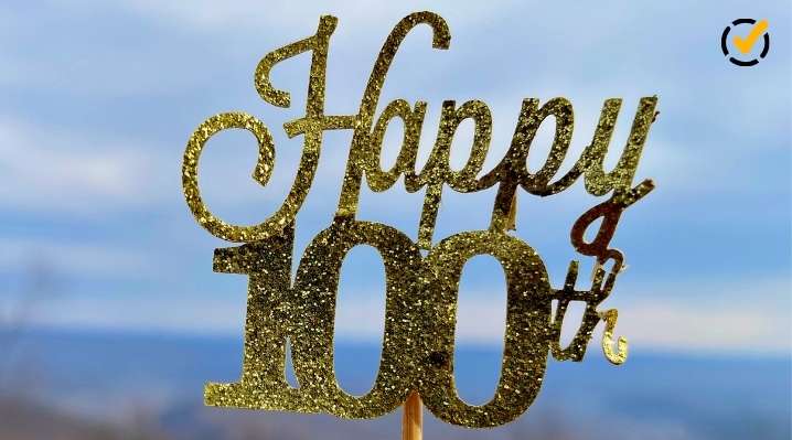 Close-up of a sparkling gold "Happy 100th" sign celebrating the 100th day of school, set against a soft bokeh background of a blue sky and distant hills. A small Formative logo is in the top right corner.