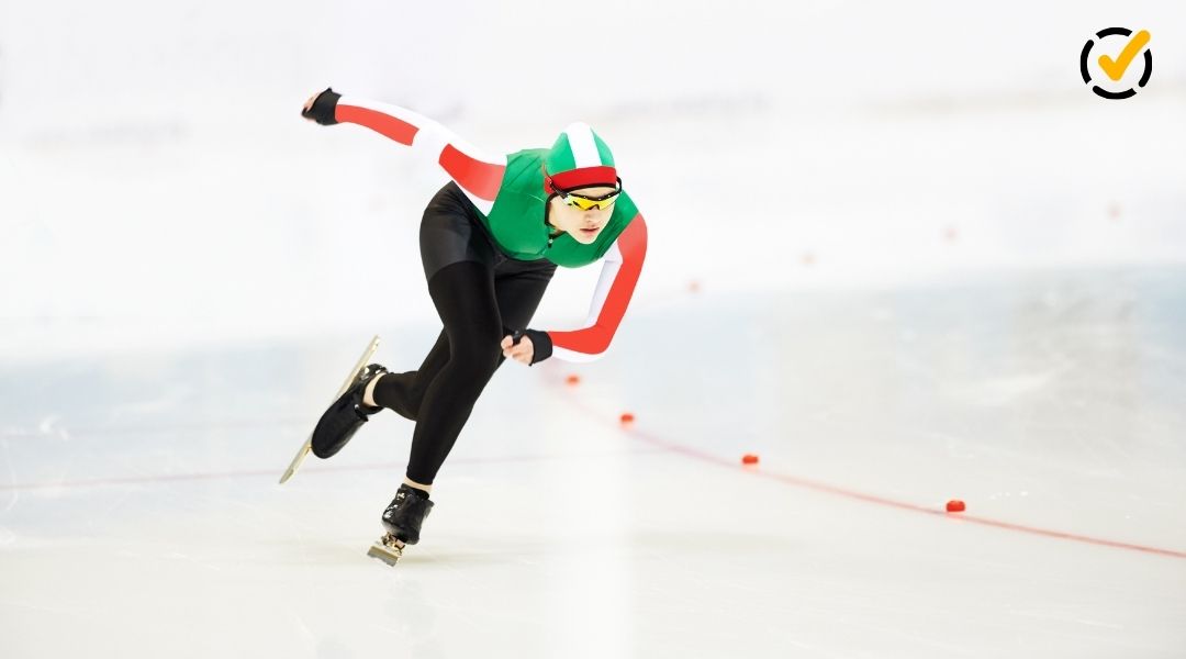 A speed skater in a green, white, and red racing suit leans into a sharp turn on an indoor ice rink. A small "check mark" logo is in the top right corner.