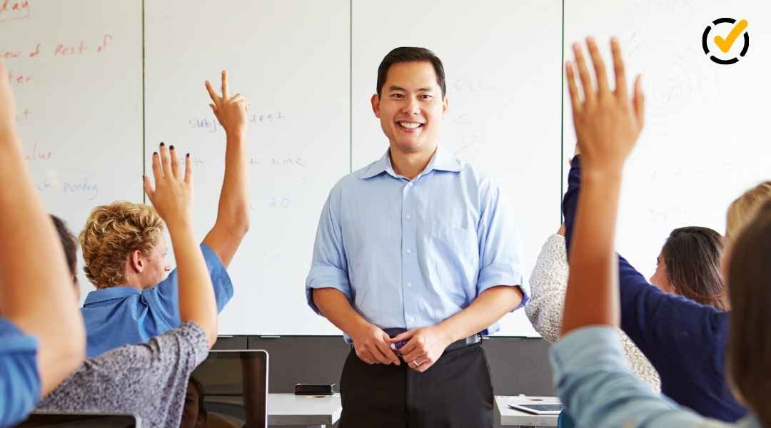 A smiling male teacher standing in front of a whiteboard as diverse students raise their hands in a bright classroom.