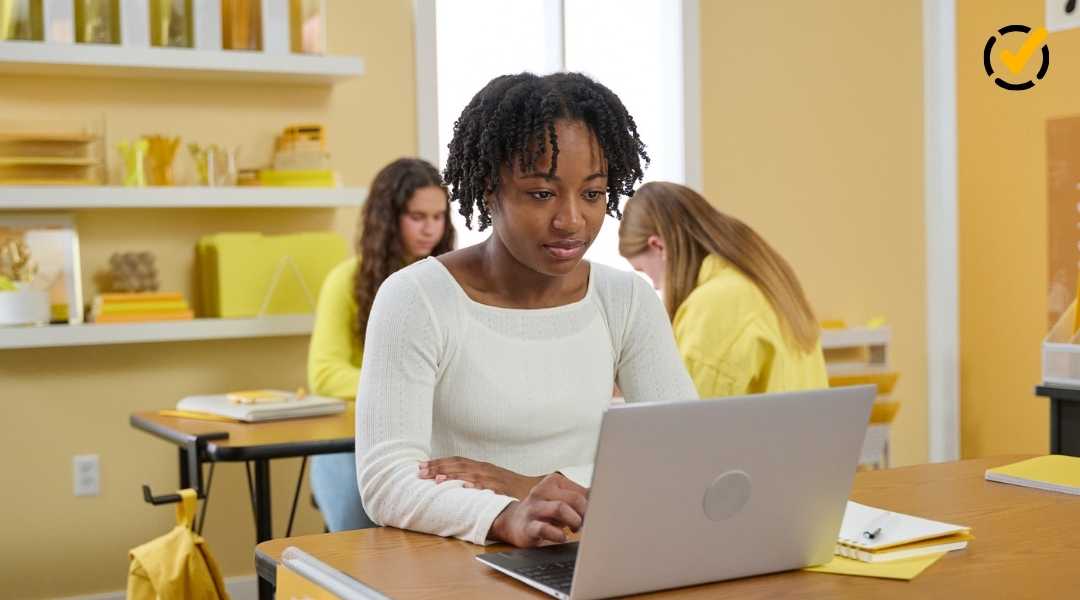 A high school student with braided hair focuses on her laptop screen while sitting at a desk in a bright, modern classroom with other students in the background.