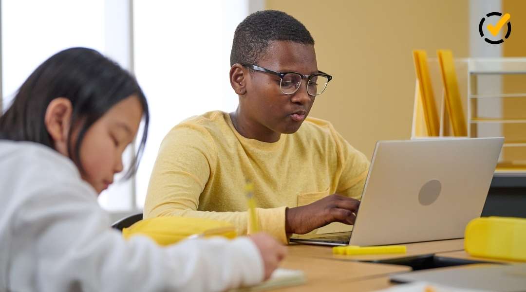 A diverse classroom setting showing a focused male student with glasses working on a laptop while a female student writes in the foreground.