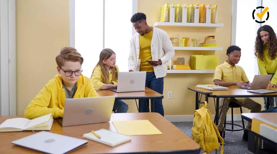 A diverse classroom of students working on laptops at individual desks while a male teacher provides one-on-one guidance. The scene uses a warm yellow color palette to reflect the Formative brand.
